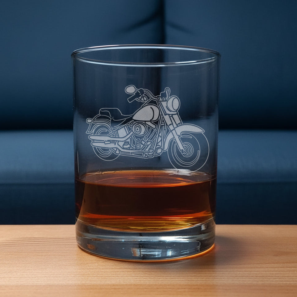 A clear glass rocks tumbler with an etched motorcycle design, partially filled with an amber-colored drink against a blue couch