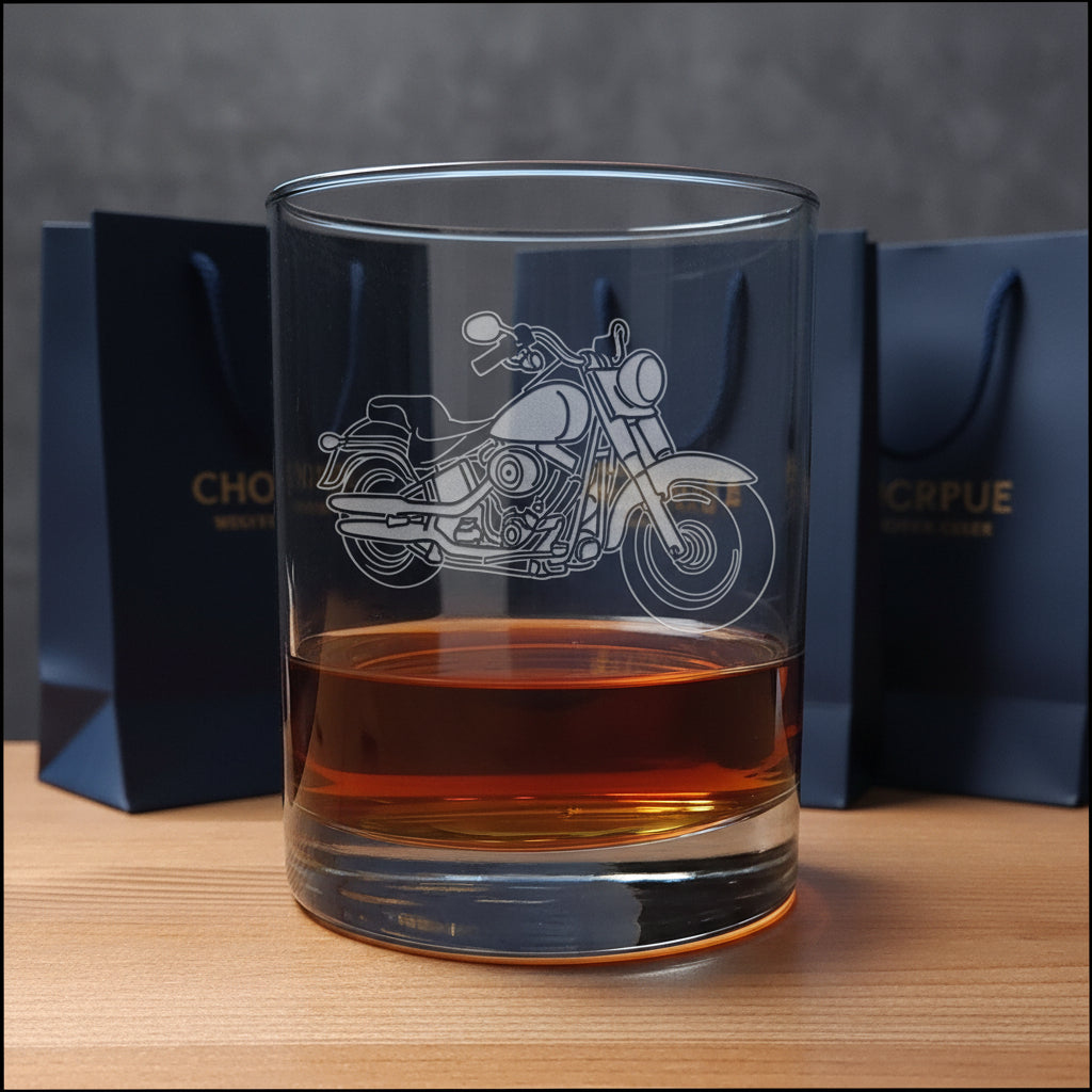 A clear glass rocks tumbler with an etched motorcycle design, partially filled with an amber-colored drink with  gift bags in the background