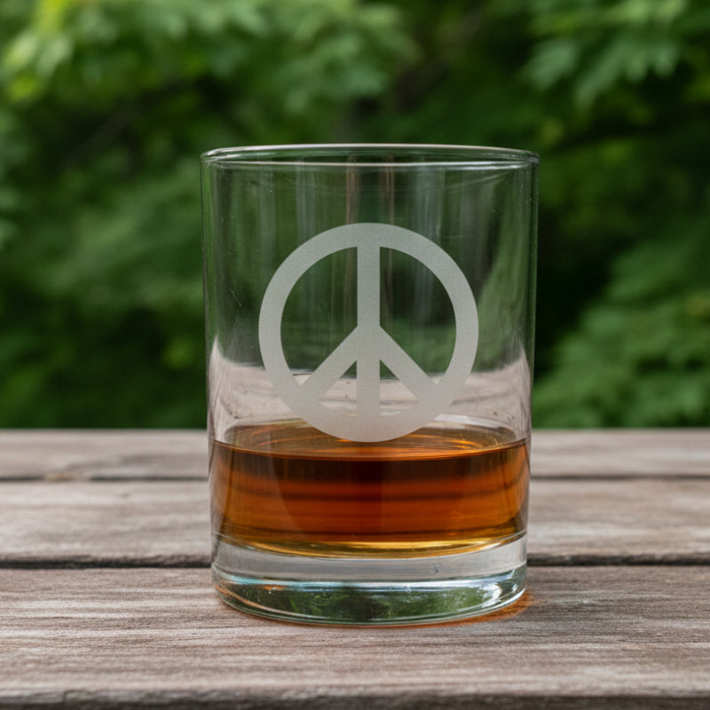 A clear rocks glass with a frosted peace sign, partially filled with amber whiskey, sitting on a light wood surface against a maple trees
