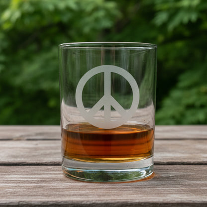 A clear rocks glass with a frosted peace sign, partially filled with amber whiskey, sitting on a light wood surface against a maple trees
