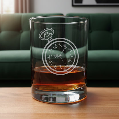 A clear rocks glass with an etched pocket watch design, partially filled with amber whiskey, sitting on a light wood surface against a green couch