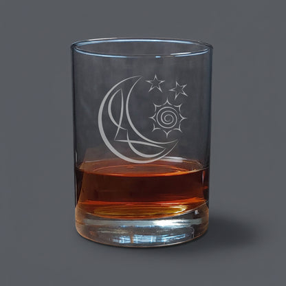 A clear glass rocks tumbler with an etched crescent moon and stars design, partially filled with amber-colored liquid, set on a wooden surface against a dark gray background.