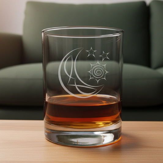 A clear glass rocks tumbler with an etched crescent moon and stars design, partially filled with amber-colored liquid, set on a wooden surface against a green couch background.