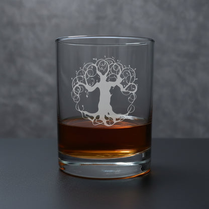 A clear glass rocks tumbler with an etched tree of life design, partially filled with amber whiskey, sitting on a light wood surface against a gray textured background.