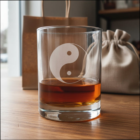 A clear rocks glass with an etched yin-yang symbol, partially filled with amber liquor, sitting on a wooden table with a brown paper bag and a beige drawstring pouch in the background.