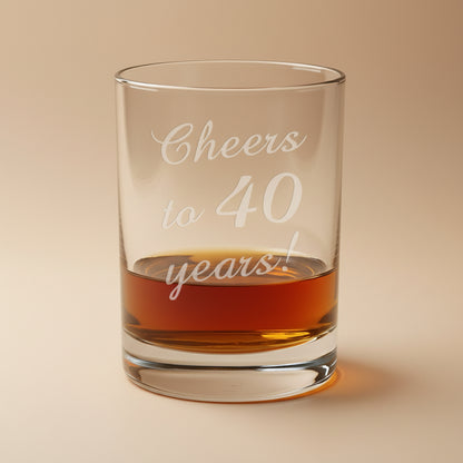 Glass with 'Cheers to 40 years!' text filled with amber liquid on a wooden surface.