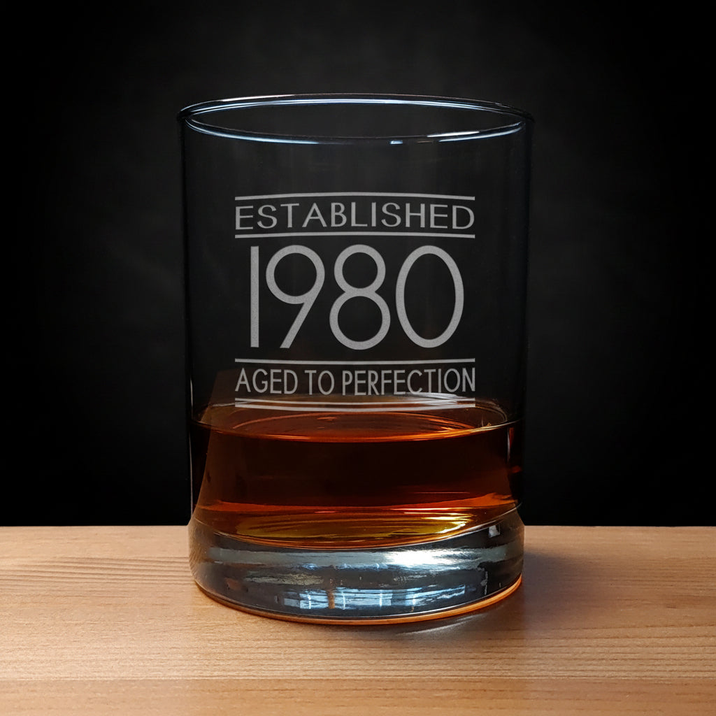 Glass tumbler with engraved text 'Established 1980 Aged to Perfection' on a wooden surface.