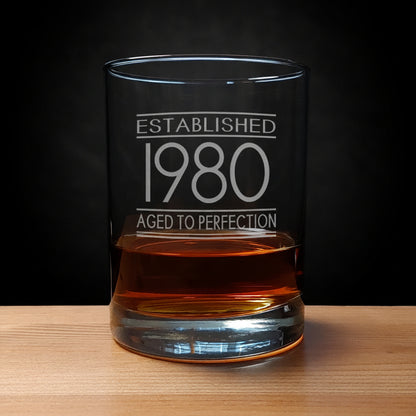 Glass tumbler with engraved text 'Established 1980 Aged to Perfection' on a wooden surface.