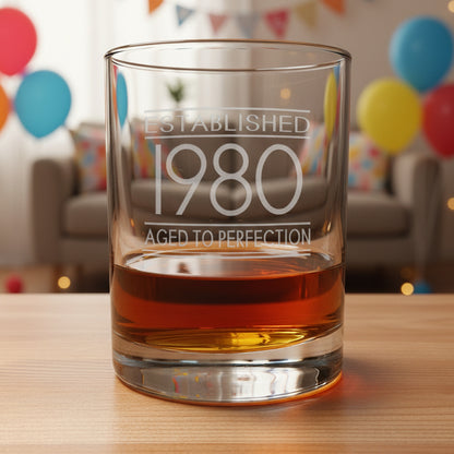 Glass tumbler with engraved text 'Established 1980 Aged to Perfection' on a wooden surface.