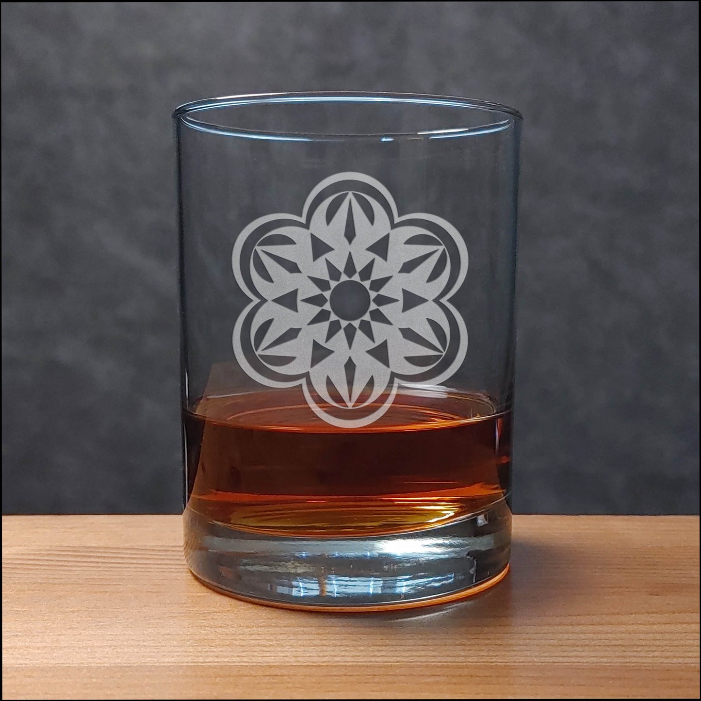 A clear rocks glass with an etched floral mandala design, partially filled with amber whiskey, set on a light wood surface against a dark gray textured background.