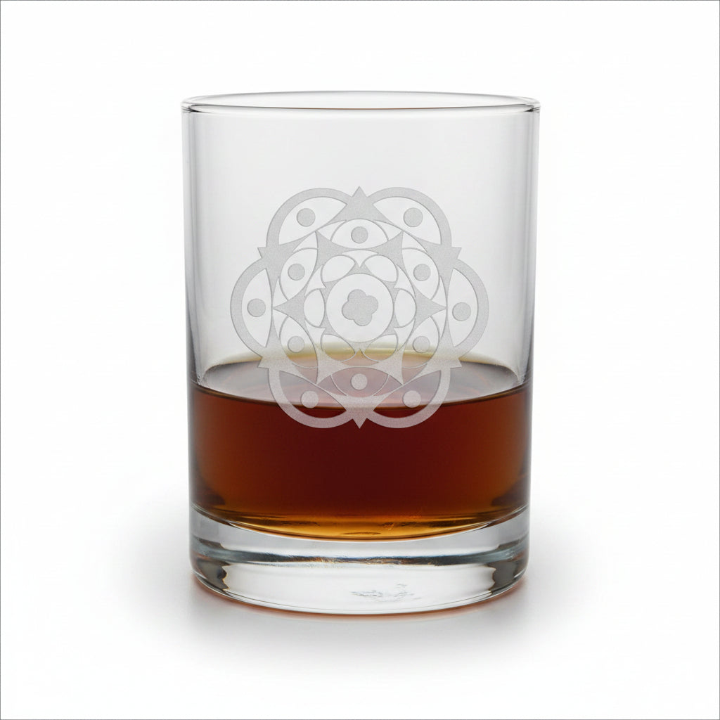 A clear glass rocks tumbler with an etched geometric mandala design, partially filled with amber-colored liquor, sitting on a light wood surface against a white background.