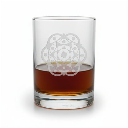 A clear glass rocks tumbler with an etched geometric mandala design, partially filled with amber-colored liquor, sitting on a light wood surface against a white background.