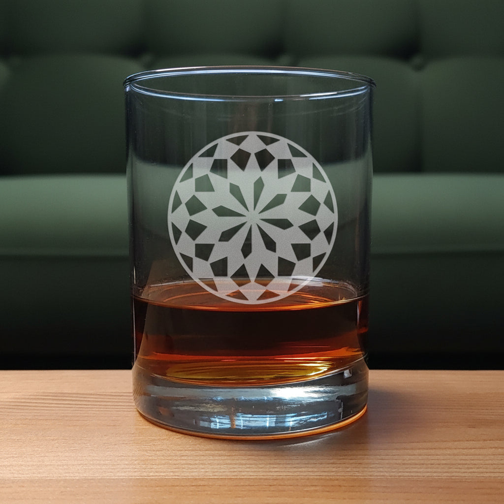 A clear rocks glass with an etched  floral mandala design, partially filled with amber whiskey, set on a light wood surface against a green couch.