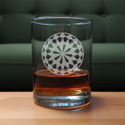 A clear rocks glass with an etched  floral mandala design, partially filled with amber whiskey, set on a light wood surface against a green couch.