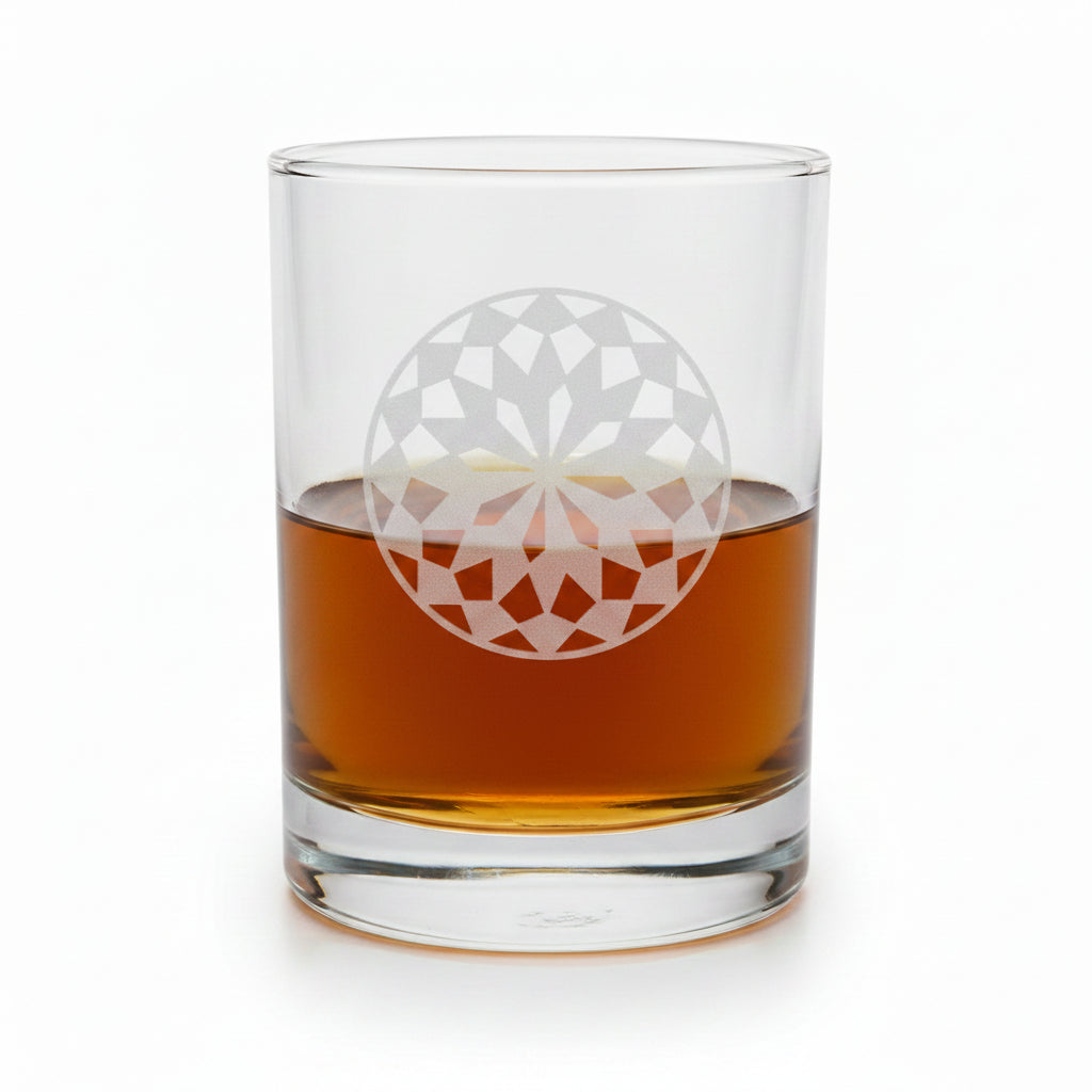 A clear rocks glass with an etched white mandala design, partially filled with amber whiskey, set on a light wood surface against a white.