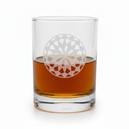 A clear rocks glass with an etched white mandala design, partially filled with amber whiskey, set on a light wood surface against a white.