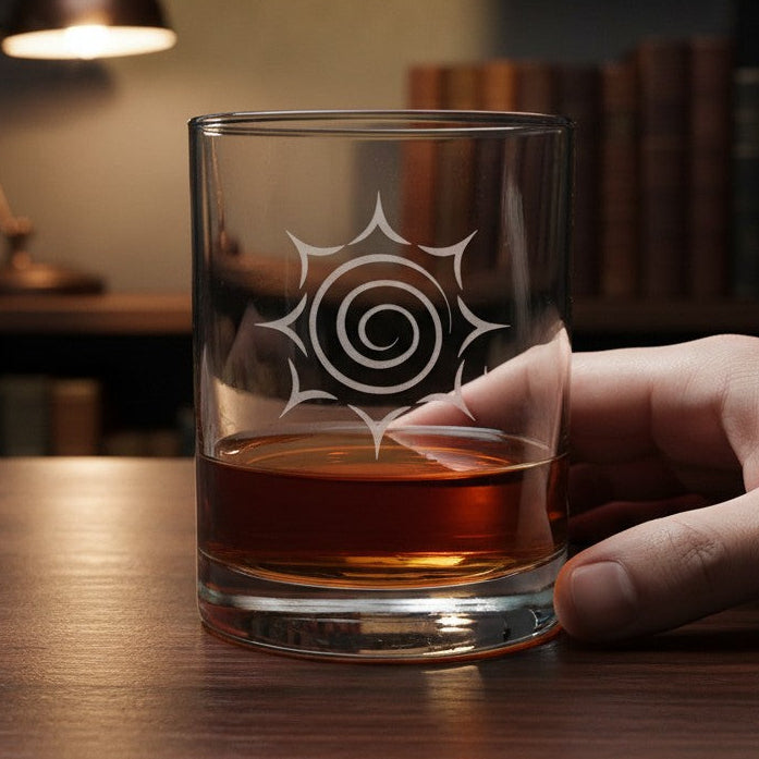 A hand holding a clear lowball glass with a white spiral sunburst design, filled with amber whiskey, set on a wooden table with a blurred bookshelf and lamp in the background.