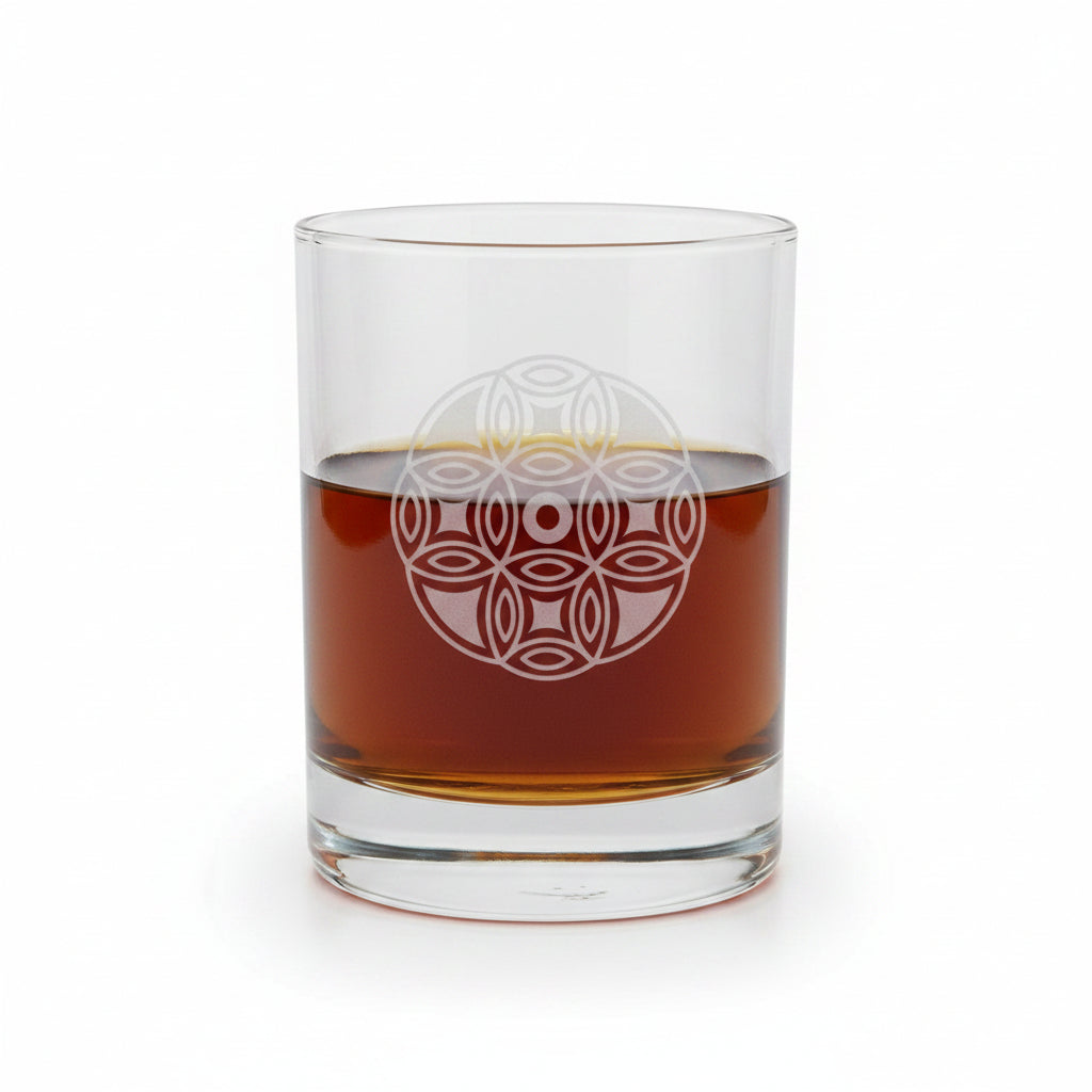 A clear glass rocks tumbler with an etched geometric mandala design, partially filled with amber whiskey, set on a wooden surface against a white background.
