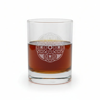 A clear glass rocks tumbler with an etched geometric mandala design, partially filled with amber whiskey, set on a wooden surface against a white background.