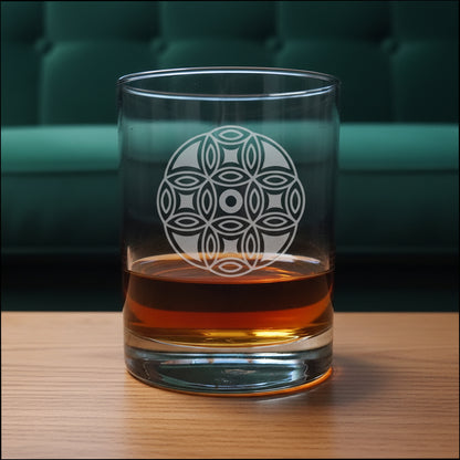 A clear glass rocks tumbler with an etched geometric flower pattern, partially filled with amber whiskey, set on a wooden surface against a green couch background.