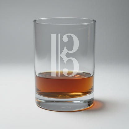 A clear glass rocks tumbler with a frosted alto clef  design, partially filled with amber whiskey, set on a light wood surface against a white background.
