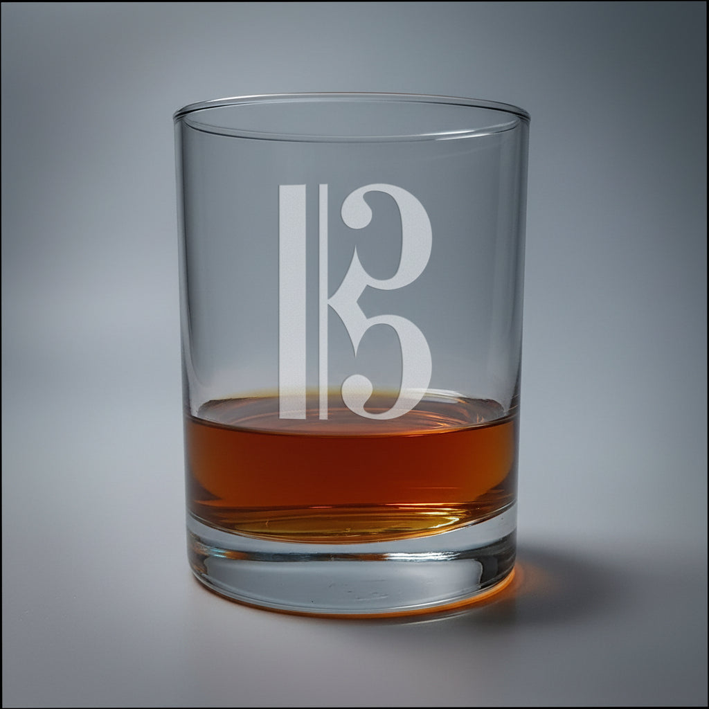 A clear glass rocks tumbler with a frosted alto clef  design, partially filled with amber whiskey, set on a light wood surface against a gray background.
