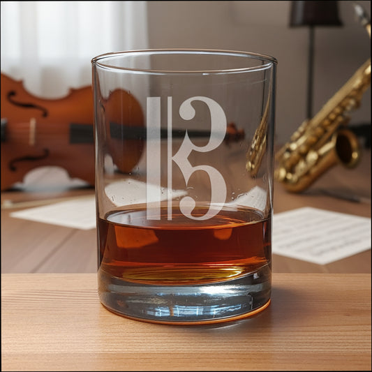 A clear glass rocks tumbler with a frosted alto clef design, partially filled with amber whiskey, set on a light wood surface against a dark gray textureda music room background.