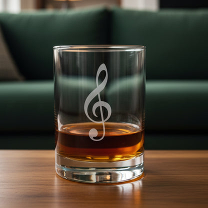 A clear glass rocks tumbler with a etched treble clef, partially filled with amber whiskey, sitting on a light wood surface against a green couch in the background.