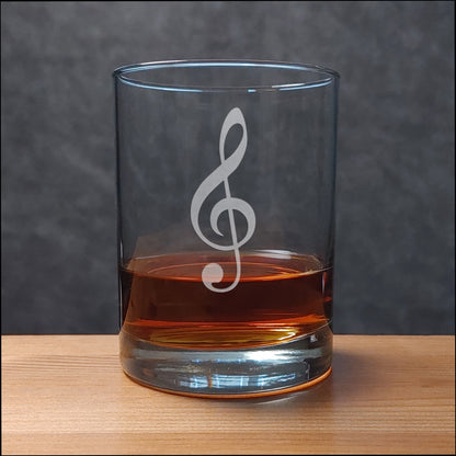 A clear glass rocks tumbler with a etched treble clef, partially filled with amber whiskey, sitting on a light wood surface against a dark gray textured background.