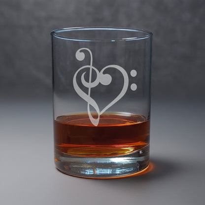 A clear glass rocks tumbler with an etched treble clef and bass clef, partially filled with amber liquor, set on a wooden surface against a gray background.