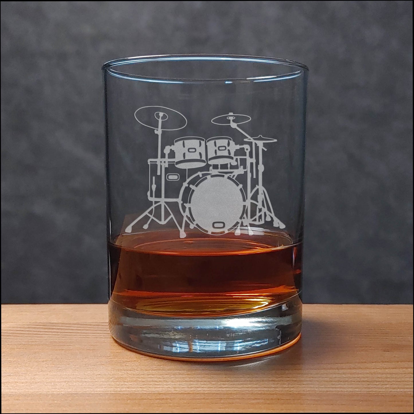 A clear rocks glass with an etched drum set design, partially filled with amber-colored liquid, set on a wooden surface against a dark gray background.