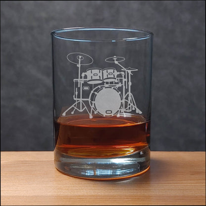 A clear rocks glass with an etched drum set design, partially filled with amber-colored liquid, set on a wooden surface against a dark gray background.