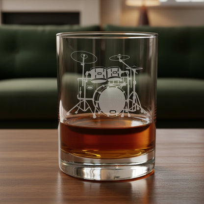 A clear rocks glass with an etched drum set design, partially filled with amber-colored liquid, set on a wooden surface against a green couch background.