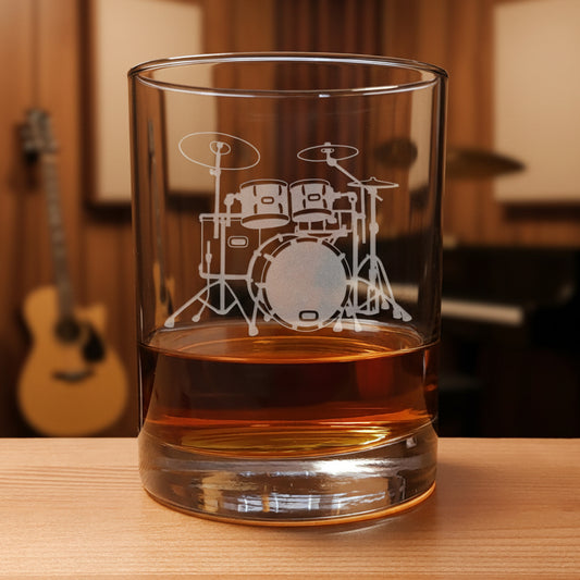 A clear rocks glass with an etched drum set design, partially filled with amber-colored liquid, set on a wooden surface against amusic room  background.
