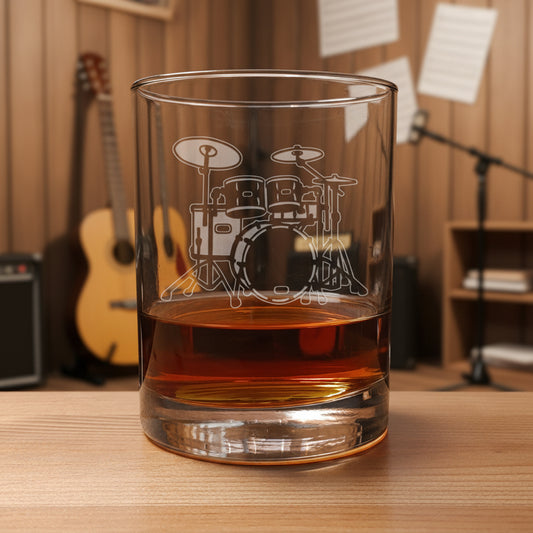 A clear rocks glass with an etched drum set design, partially filled with amber-colored liquid, set on a wooden surface against amusic room  background.