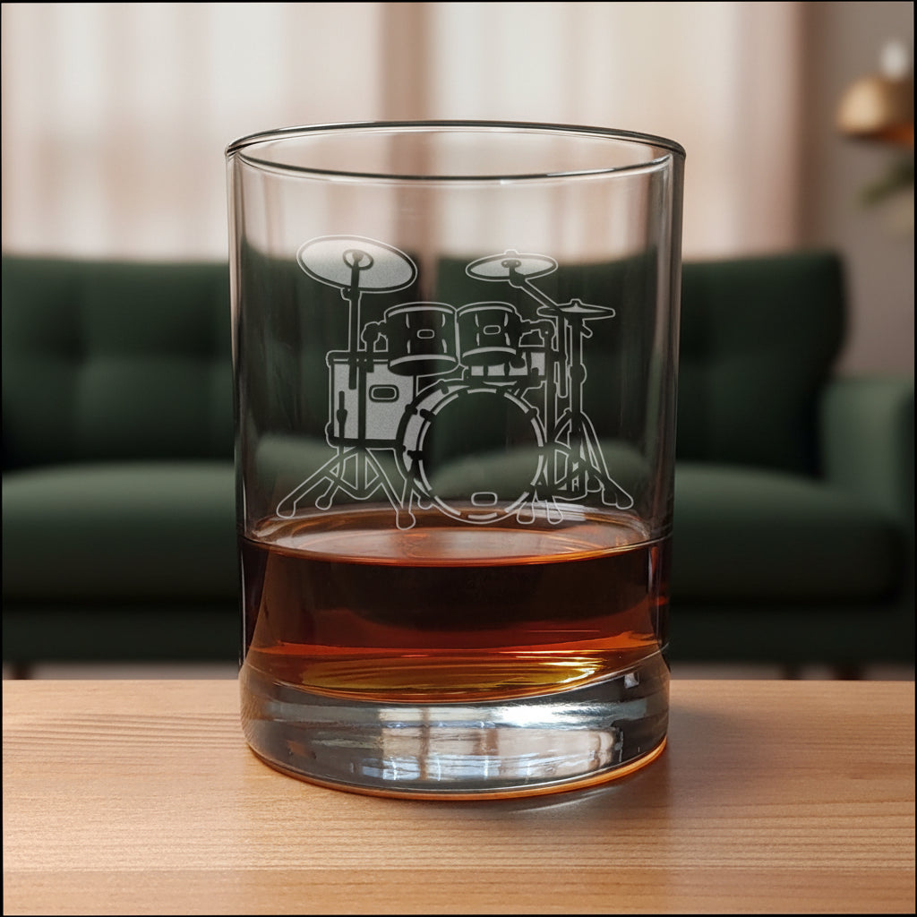 A clear rocks glass with an etched drum set design, partially filled with amber-colored liquid, set on a wooden surface against a green couch background.