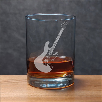A clear rocks glass with an etched electric guitar design, partially filled with an amber liquid, set on a wooden surface against a dark gray textured background.