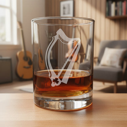 A clear glass rocks tumbler with an etched harp design and shamrock detail, partially filled with amber whiskey, set on a wooden surface the background of a music room.