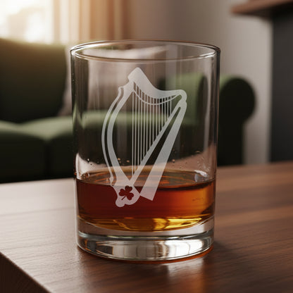 A clear glass rocks tumbler with an etched Irish harp design, partially filled with amber whiskey, resting on a light wood surface against 