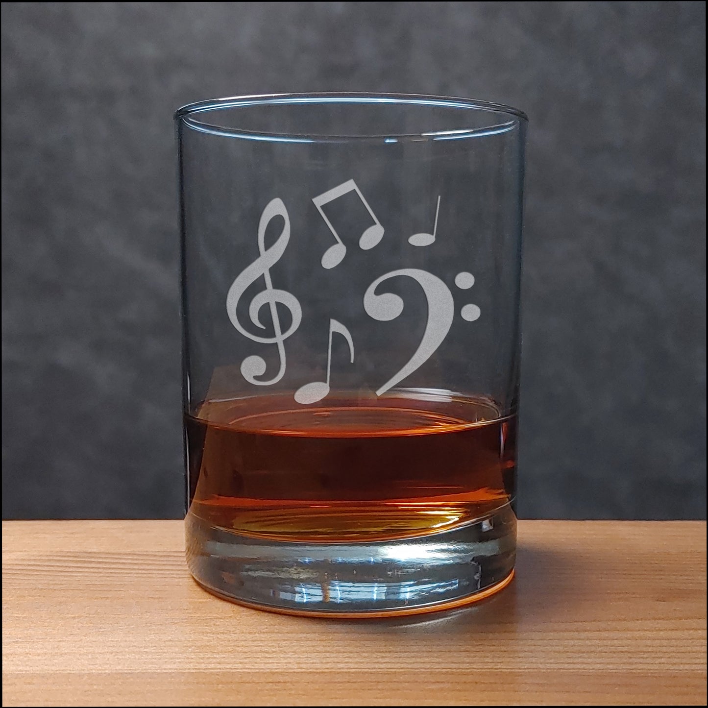 A clear glass rocks tumbler with white etched musical notes (treble clef, bass clef, and eighth notes) sitting on a light wood surface against a dark gray textured background. The glass is partially filled with an amber-colored liquor.