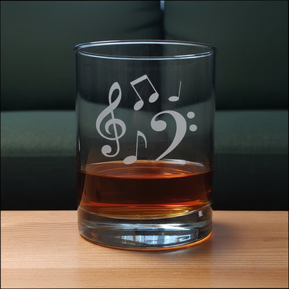 A clear glass rocks tumbler with  etched musical notes (treble clef, bass clef, and eighth notes) sitting on a light wood surface against a green couch  in the  background. The glass is partially filled with an amber-colored liquor.