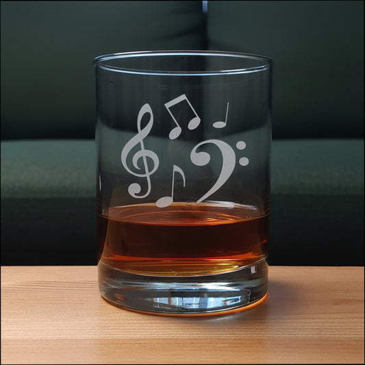 A clear glass rocks tumbler with  etched musical notes (treble clef, bass clef, and eighth notes) sitting on a light wood surface against a green couch  in the  background. The glass is partially filled with an amber-colored liquor.