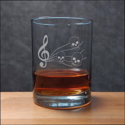 A clear glass rocks tumbler with an etched treble clef and three skulls, partially filled with an amber drink, set on a wooden surface against a dark gray background.