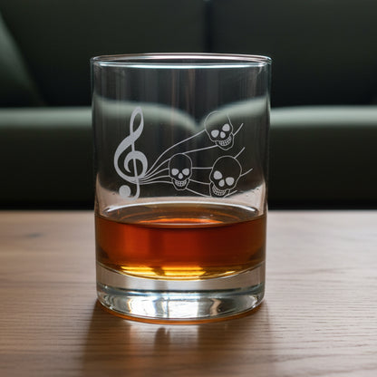 A clear glass rocks tumbler with an etched treble clef and three skulls, partially filled with an amber drink, set on a wooden surface 