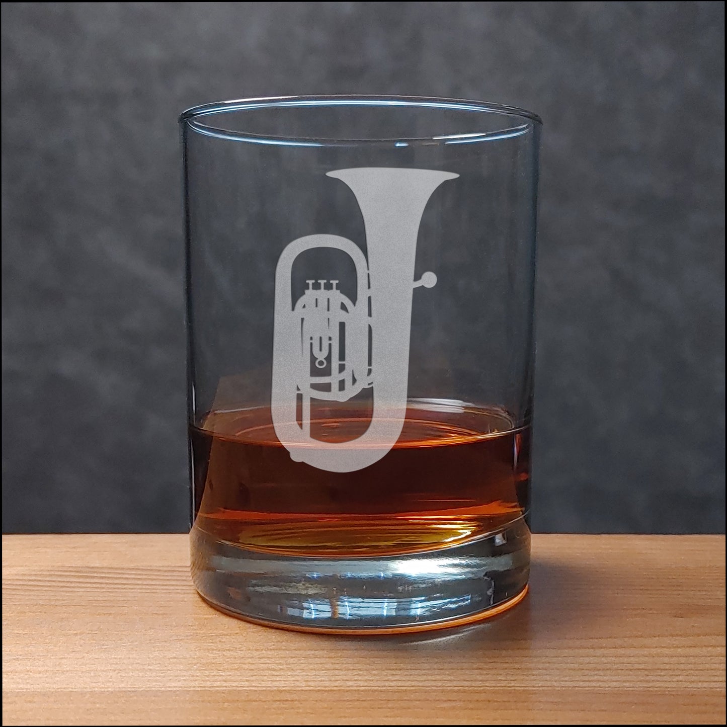 A clear glass rocks tumbler with a white etched trumpet design, partially filled with amber whiskey, set on a light wood surface against a dark gray textured background.