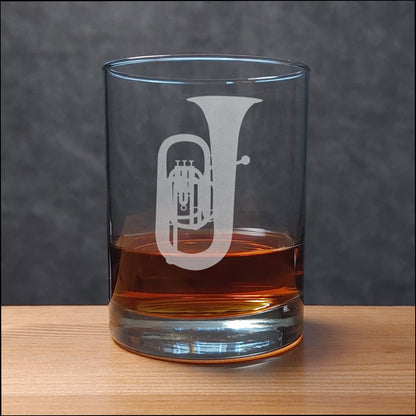 A clear glass rocks tumbler with a white etched trumpet design, partially filled with amber whiskey, set on a light wood surface against a dark gray textured background.