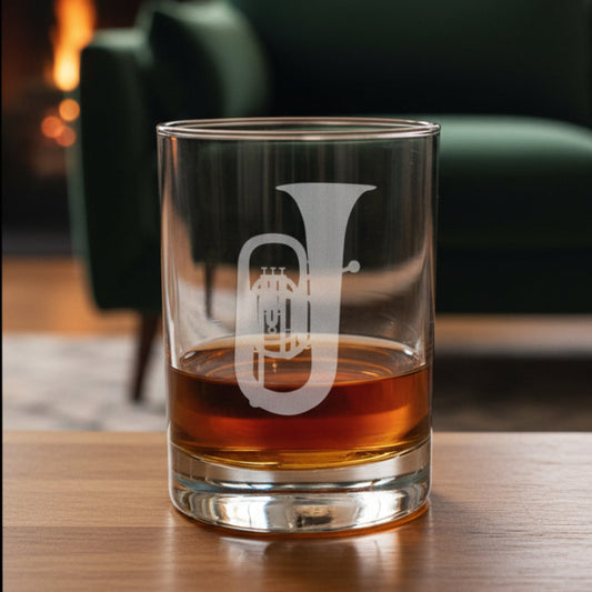 A clear rocks glass with an etched tuba design, partially filled with amber-colored liquid, set on a wooden surface against a green couch background.