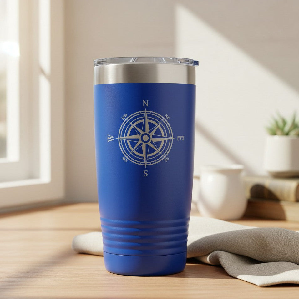 A royal blue insulated tumbler with a silver stainless steel rim and a white compass rose graphic on the front, set on a wooden table in a bright, modern room.