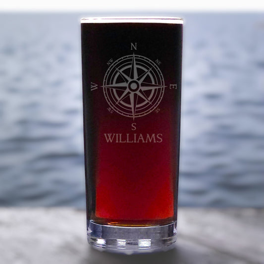 Pint glass with compass rose and 'Williams' engraving, filled with red liquid, on a blurred water background.