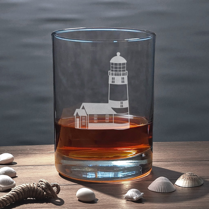 Glass with lighthouse design filled with amber liquid on a wooden surface with shells.
